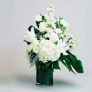 Tall vase arrangement in all white flowers accented with tropical greens.