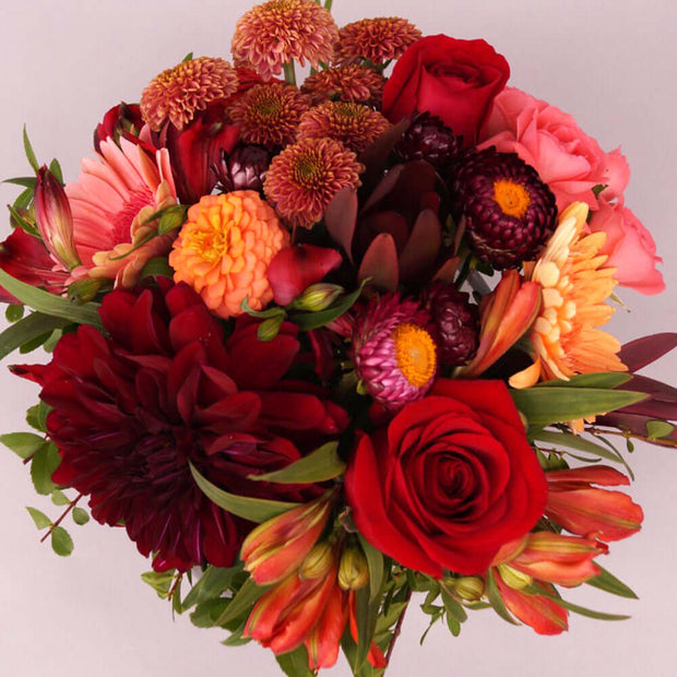 All-around flowers in red, burgundy, pink, peach, and coral, accented with assorted greenery designed in our low round clear vase.